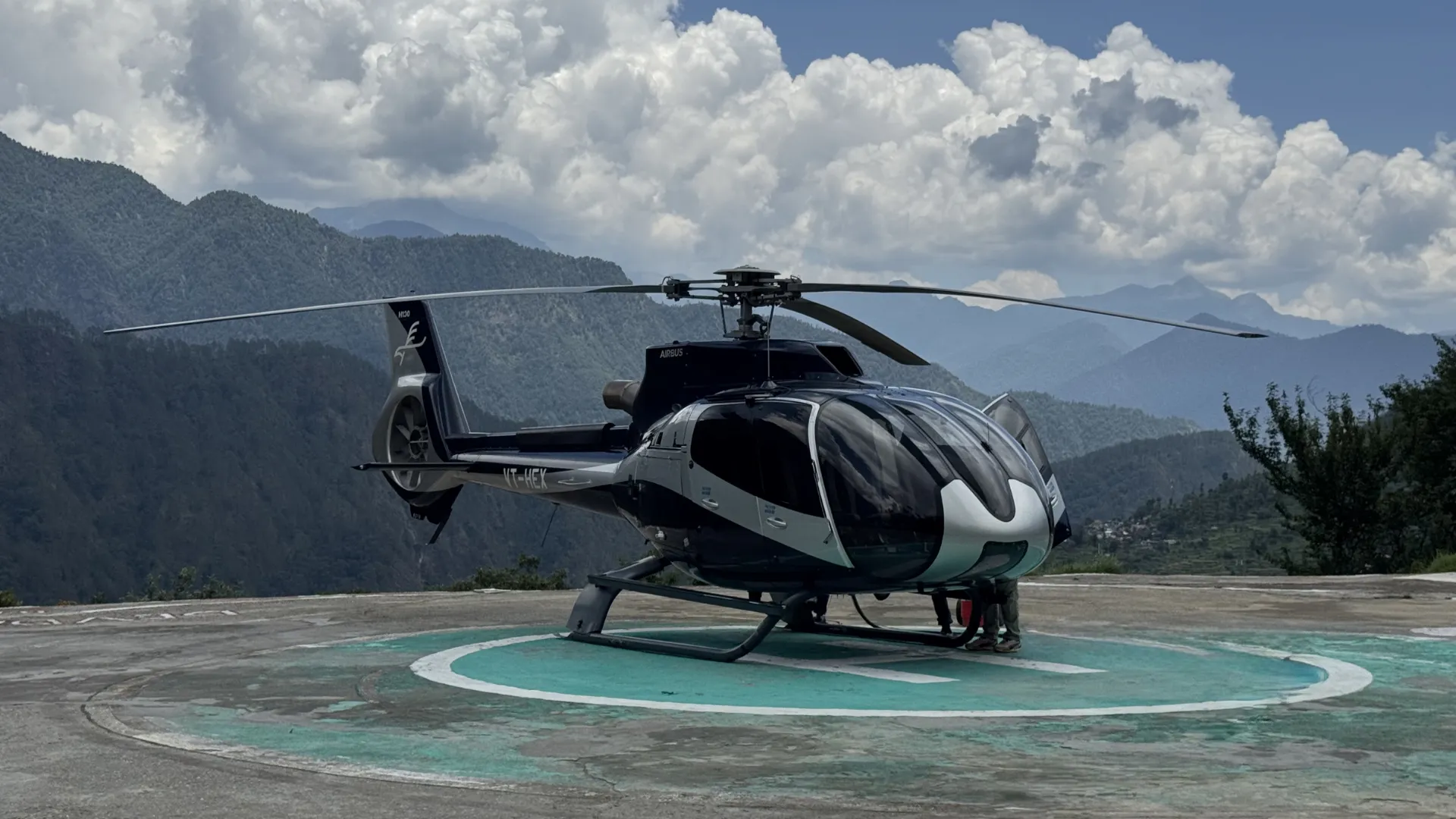 A black and silver helicopter parked on a mountain helipad with green hills and a cloudy sky in the background Captured Through chardham Package for Chardham Yatra By Helicopter Booking.
